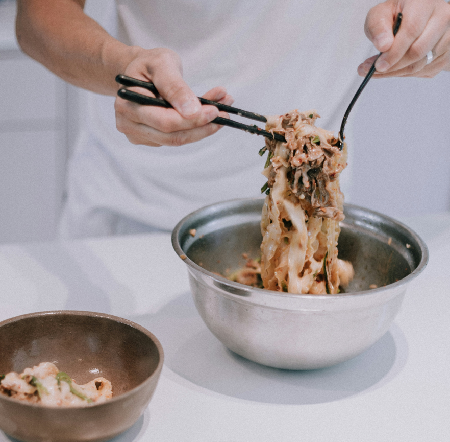 A person using chopsticks and a fork to lift noodles with beef from a metal bowl, showcasing a freshly prepared noodle dish.