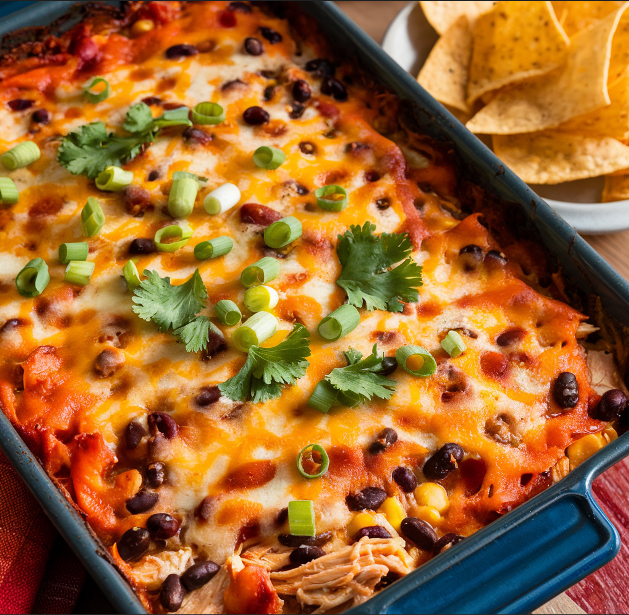 A flat lay of ingredients for Enchilada Chicken Casserole, including shredded chicken, enchilada sauce, corn tortillas, shredded cheese, black beans, green chilies, frozen corn, and chopped cilantro, arranged on a wooden surface with a baking dish and festive decorations.